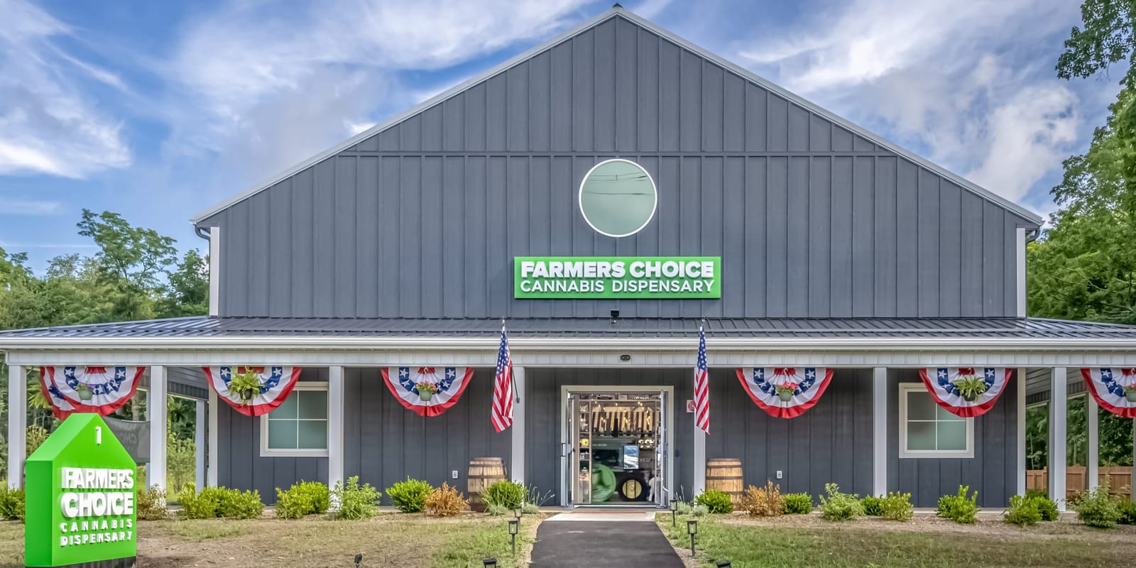 The Farmers Choice New Paltz barn seen from its front lawn, the green Farmers Choice sign beside the path, bunting and flags along the white-columned porch under a summer sky
