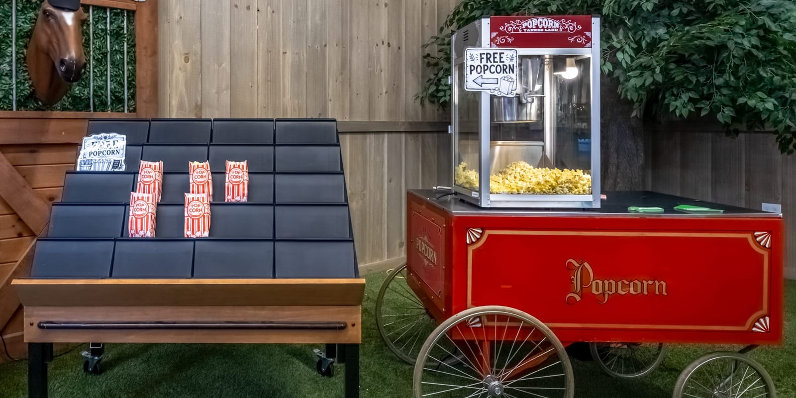 A red vintage popcorn cart with a popcorn machine on top beside a black display table of popcorn bags, against the plank walls inside the Farmers Choice New Paltz barn