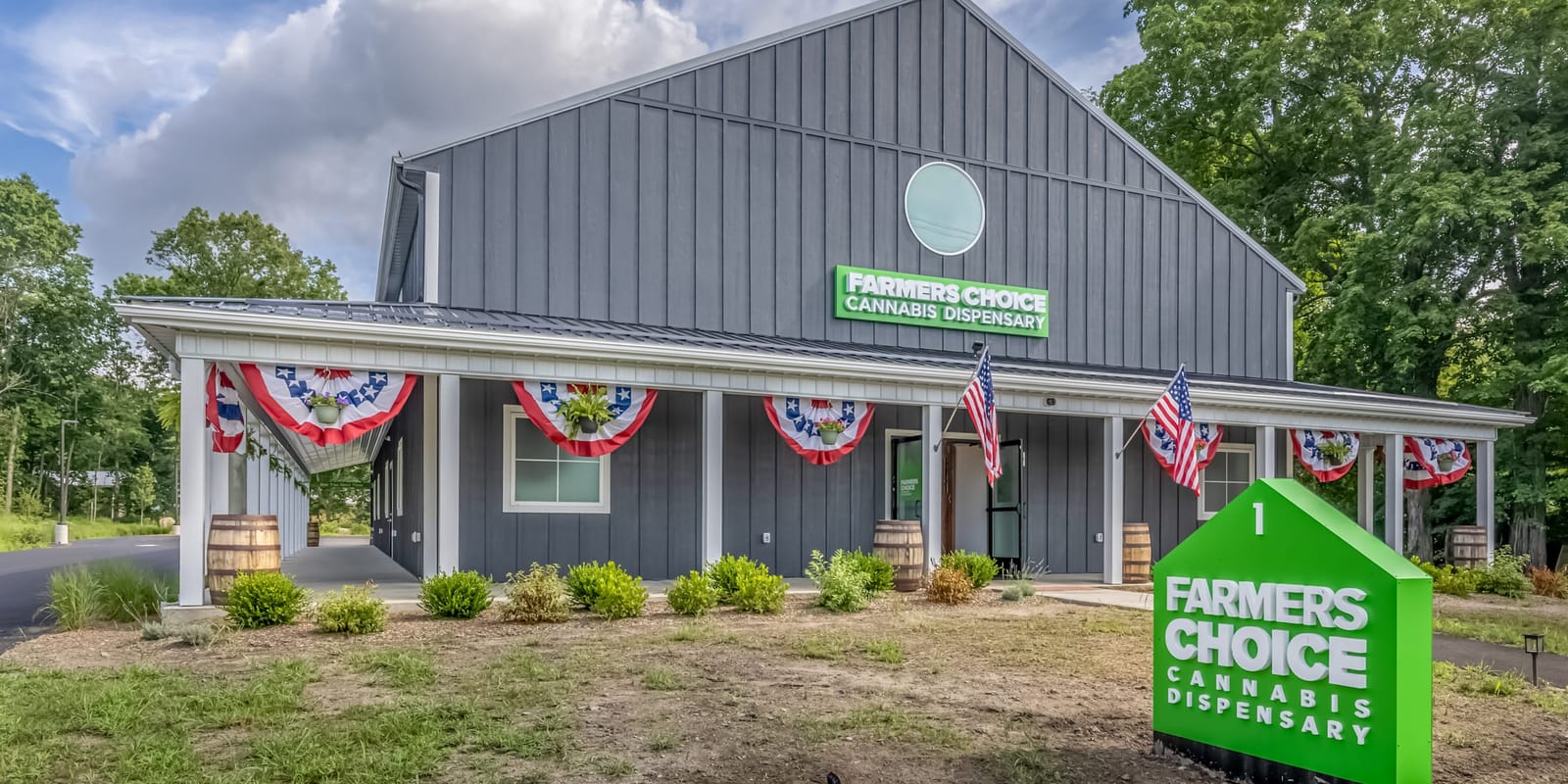 The Farmers Choice New Paltz barn under a bright cloudy sky, the green house-shaped Farmers Choice sign in the foreground and bunting along the porch