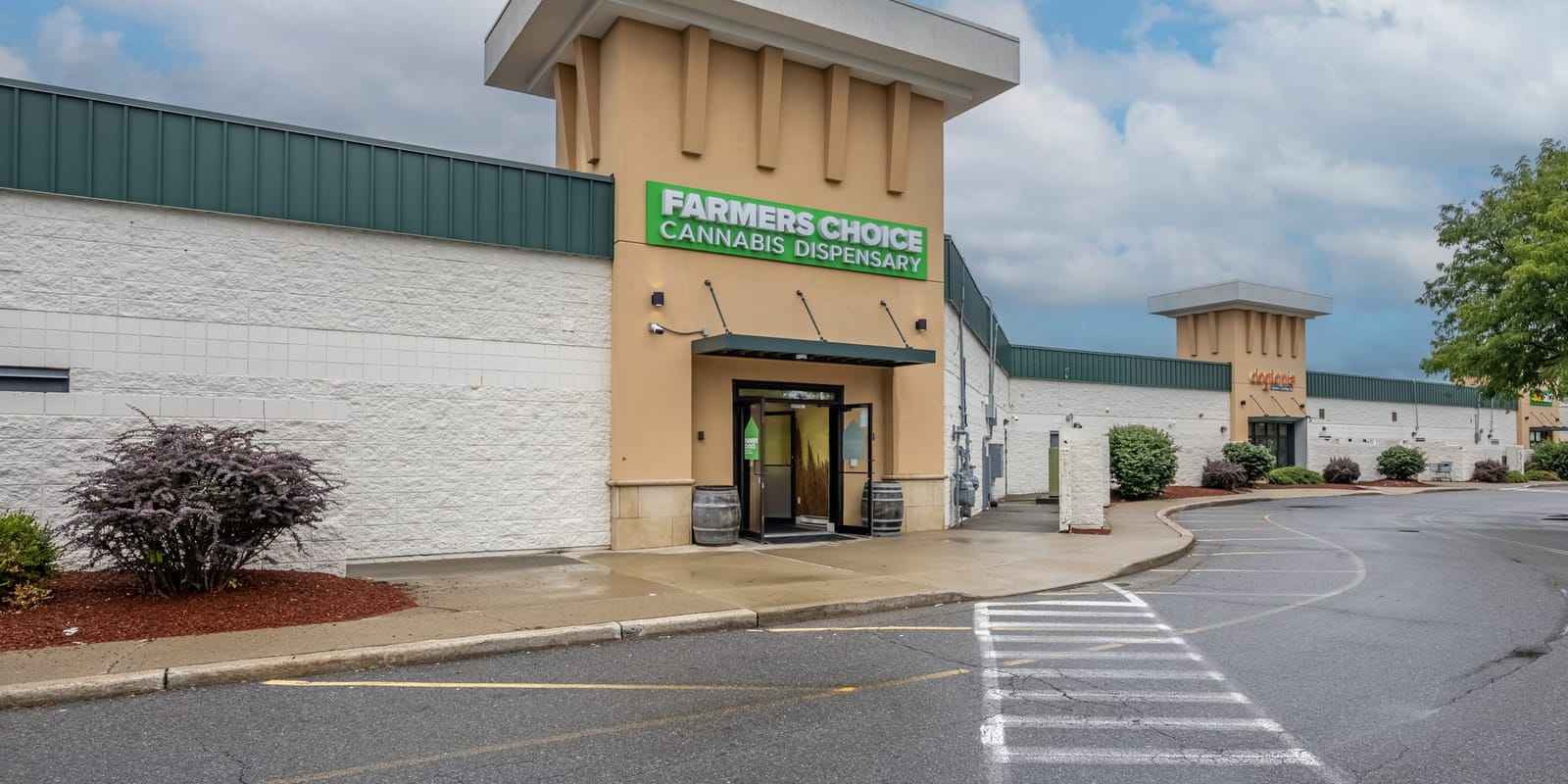 The Farmers Choice Fishkill entrance tower from the corner of its parking lot, a green sign under a cloudy sky and shrubs along the wall