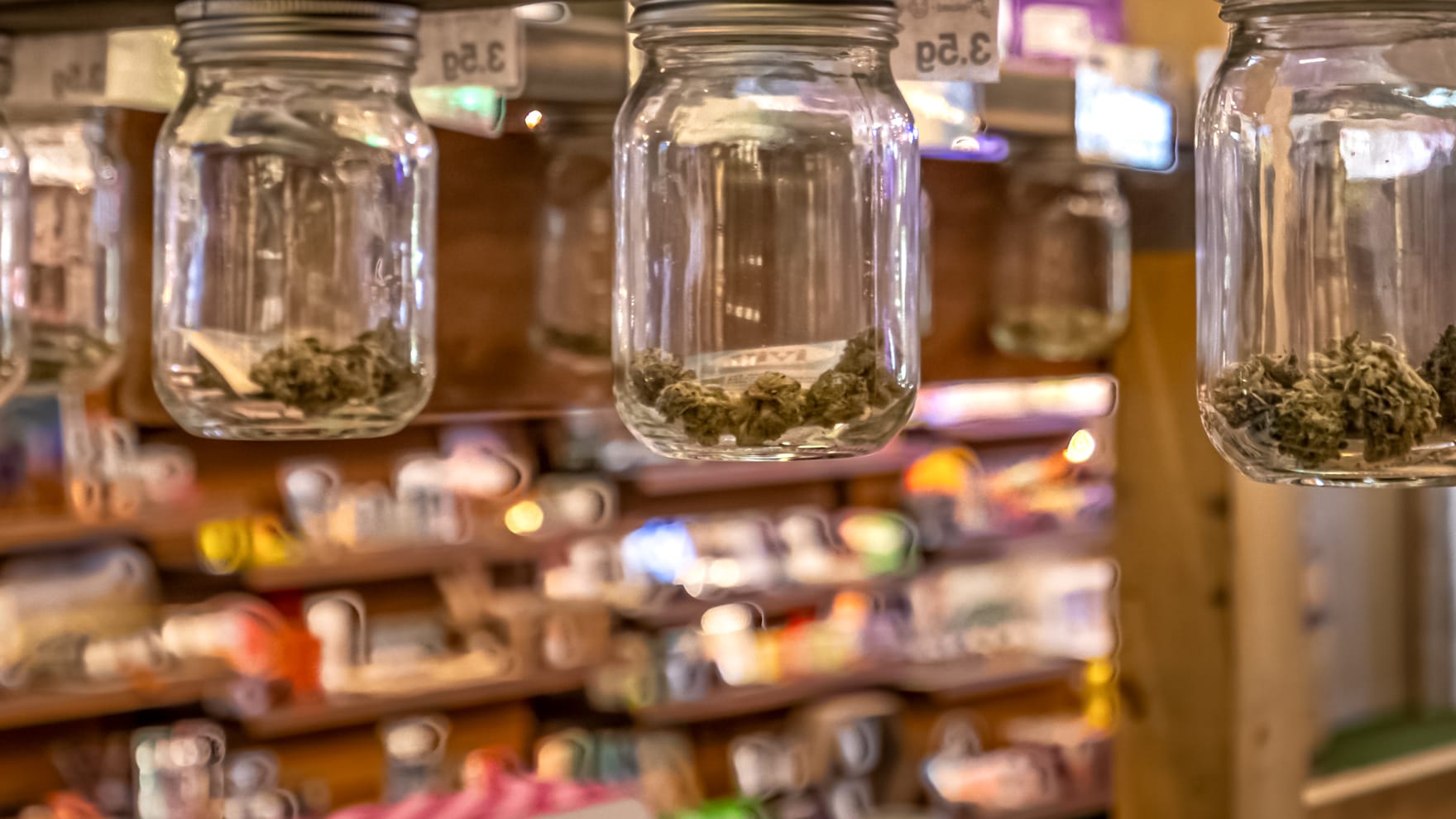 Glass mason jars of cannabis flower hanging under a wooden shelf at the Farmers Choice New Paltz counter, the product wall soft behind them