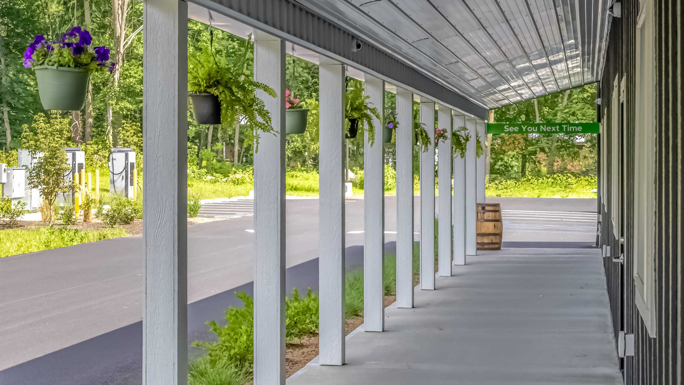 The covered porch along the Farmers Choice New Paltz barn, white columns, hanging ferns and petunias, the EV chargers and parking beyond